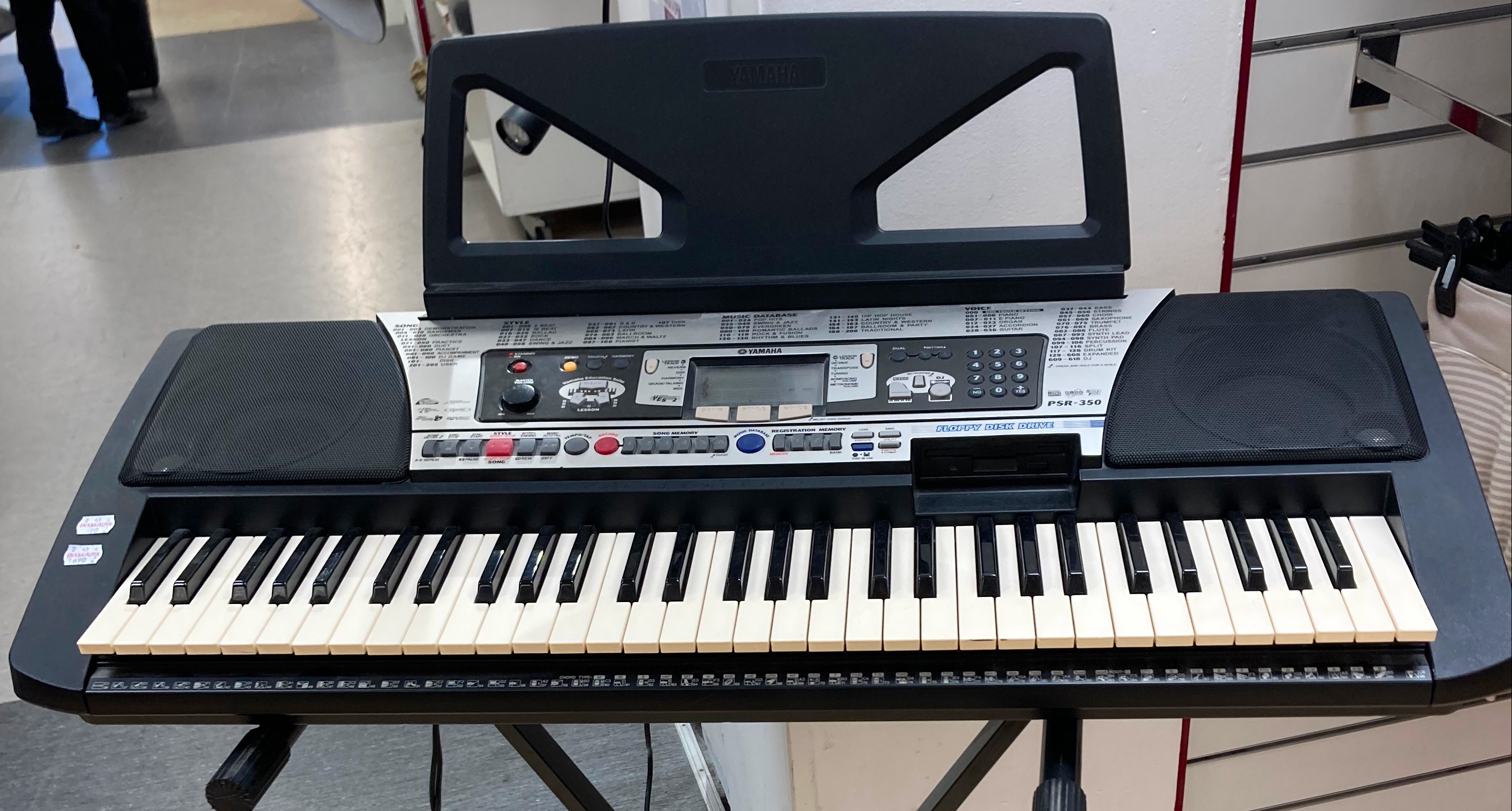 Photo of a Yamaha PortaTone PSR-350 sitting on a keyboard stand. It looks like any other large portable keyboard instrument in its class. It has big speakers on either side, 61 fairly large plastic keys, a floppy disk drive, and a music rest. It also has large amounts of text printed on the plastic listing different kinds of instrument sounds, built-in songs, styles, and “Music Database” entries. There are many rubber buttons for controlling (e.g. starting and stopping) song and auto-accompaniment playback, for controlling the “Song Memory” and “Registration Memory”, for accessing the settings and the “Lesson” feature, for entering numbers, and so on. There is a dedicated “Portable Grand” button and a “DJ” button. There is a large volume knob, a power switch, and a dedicated “Demo” button. In the centre is an LCD that is relatively small in relation to the unit, but still larger than the LCD on any half-rack unit, and which uses fixed LCD segments rather than dot-matrix graphics. The LCD has a treble clef, bass clef and stave lines permanently superimposed on it. The LCD is not currently illuminated because the keyboard is switched off. The plastic on the unit features a great many logos that advertise various features, but only the “General MIDI” one would be recognisable to someone without deep familiarity with the Yamaha PSR series.