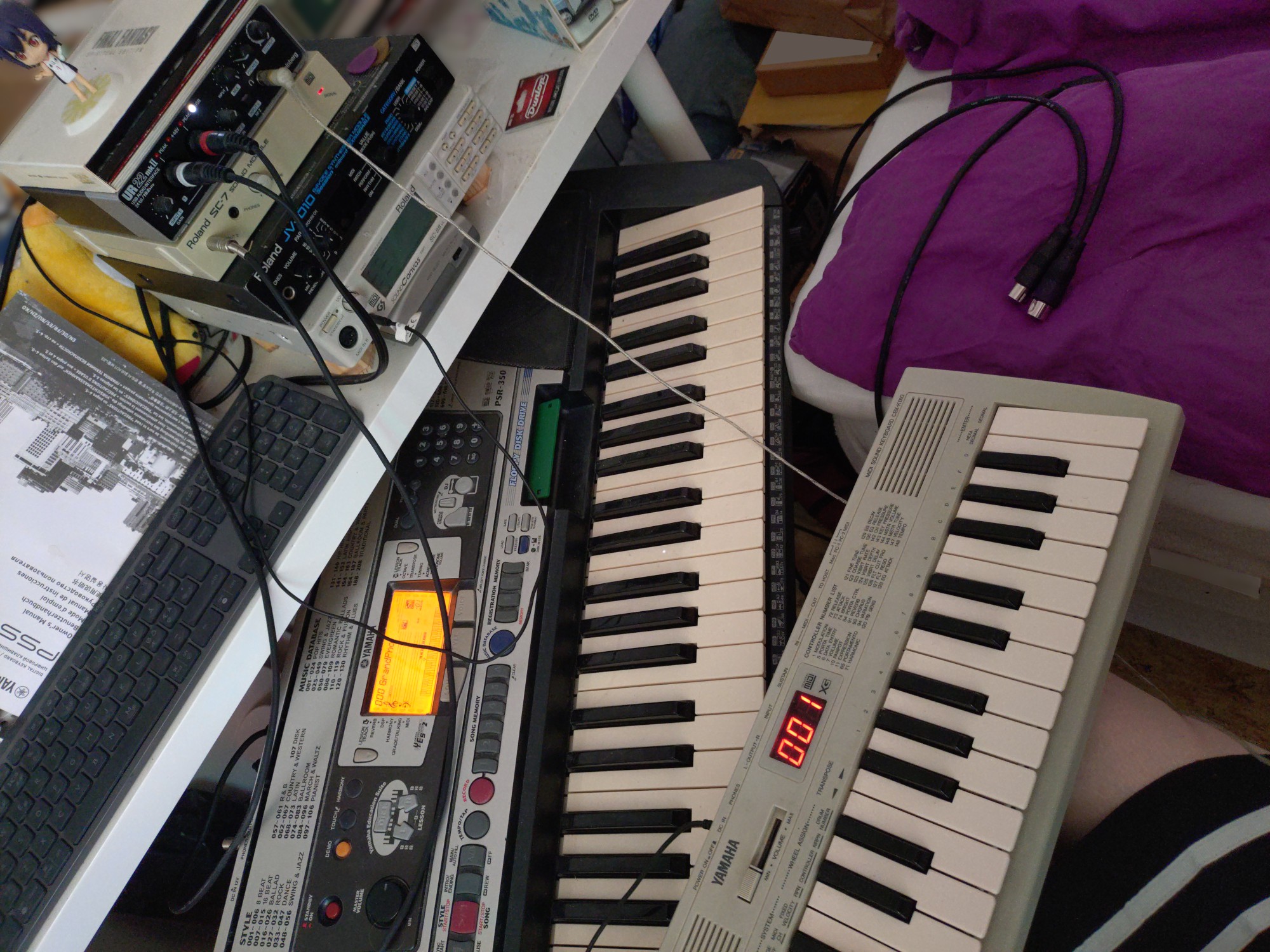 A photo of a part of someone's bedroom, taken from something like their eye level, sitting down. In the top-left part of the image, a desk can be seen, which has among other things a computer keyboard and the Yamaha PSS-A50 manual on it, but the main focus here is a stack of MIDI and audio equipment, from bottom to top: a Yamaha QY70, a Roland SC-88VL, a Roland JV-1010, a Roland SC-7, a Steinberg UR22mkII audio interface, a DVD of “Final Fantasy: The Spirits Within”, and a tiny chibi figurine of the Kanbaru Suruga from the Monogatari series. Various cables can be seen connecting some of the devices to eachother, which hang over large parts of the photo, and both the UR22mkII and SC-7 are powered on. Below the desk, a powered-on Yamaha PSR-350 can be seen, poking out somewhat awkwardly, and with its screen glowing in brilliant orange. On top of the PSR-350, but actually lying in someone's lap, is a Yamaha CBX-K1XG, also powered on. The person is wearing a relatively short skirt with a black-and-white striped pattern. The corner of a bed is visible to the right of the desk and the keyboards, and a traditional MIDI cable that is not connected to anything is lying on it. The overall impression is neither fully orderly nor fully chaotic. Some Dunlop guitar picks and their packaging can also be seen.