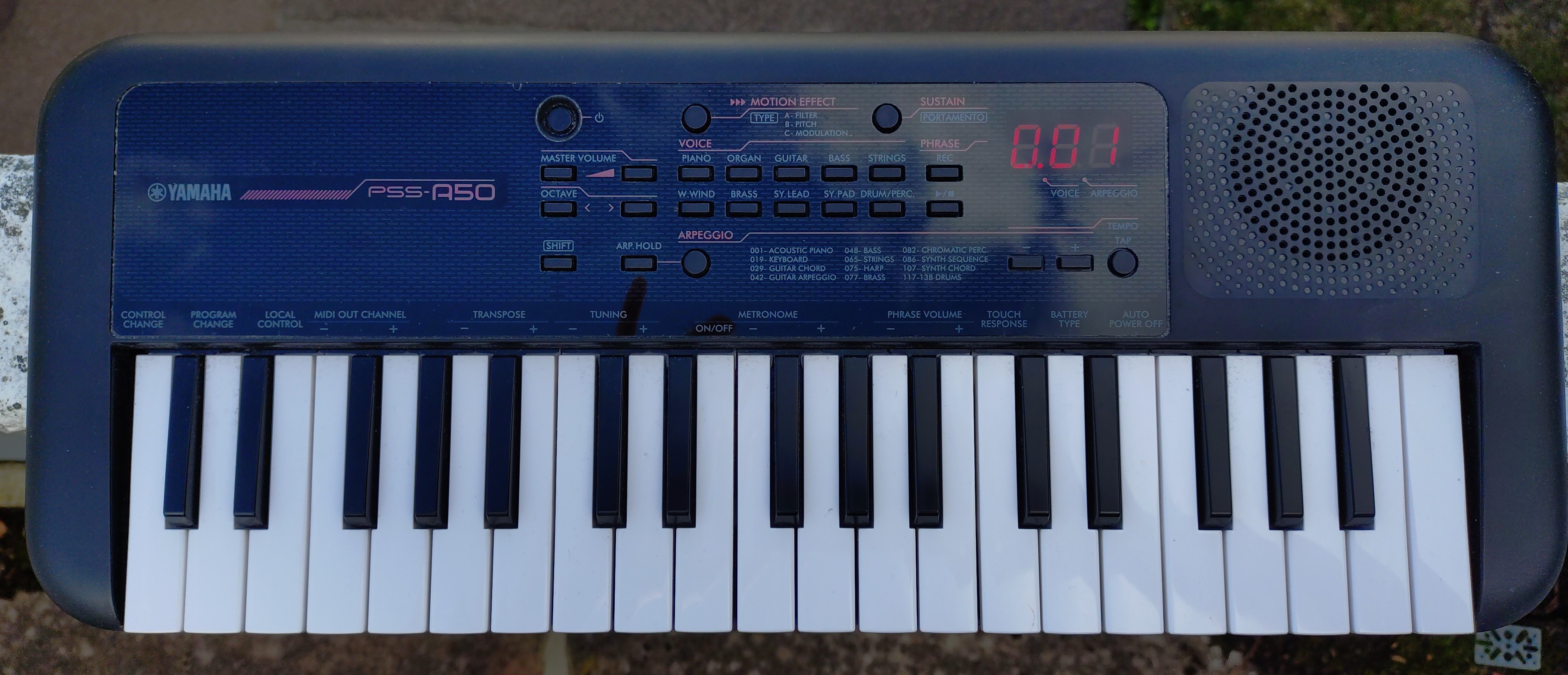 Photo of a Yamaha PSS-A50. This is a small, portable, battery-powered keyboard with 37 mini keys. The enclosure is made from black plastic, with rounded corners on all sides, and the top-left and top-right corners being almost spherical. It has a single, large speaker grille, a red-on-black three-digit seven-segment LED display, and a selection of buttons for selecting different categories of “voices” (for example, “Piano”, “Bass” and “Brass”), for adjusting the “Master Volume” and “Octave”, for activating the “Arpeggio”, “Motion Effect” and “Sustain“, for setting the “Tempo”, and for recording “Phrases”. There are also various other features (which generally have something to do with MIDI) indicated with labels above the keyboard keys. The photo has obviously been taken outdoors in natural light, with shiny part of the plastic reflecting the blue of the sky and the clouds.