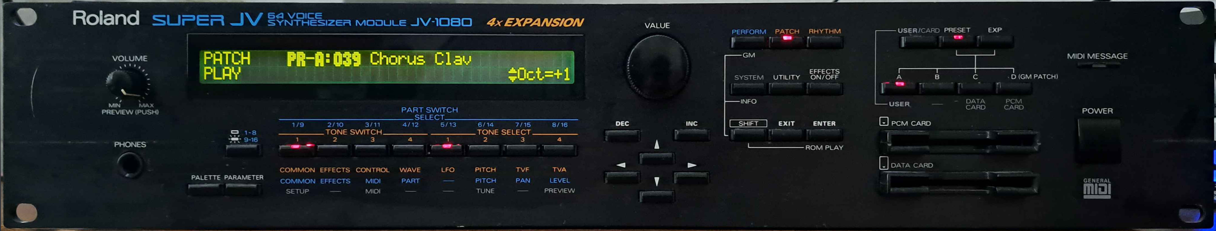 Photo of a Roland “Super JV 64 Voice Synthesizer Module JV-1080”, that proudly claims it has “4× EXPANSION”. This is a huge, full-width rack-mounted unit (2U height) with a black metal enclosure. It has a big two-line green-on-dark-green character LCD, many buttons for selecting and editing patches, a big encoder wheel, a “PCM Card” slot, a “Data Card” slot, a quarter-inch “Phones” jack, and a prominent “General MIDI” logo, among other things.