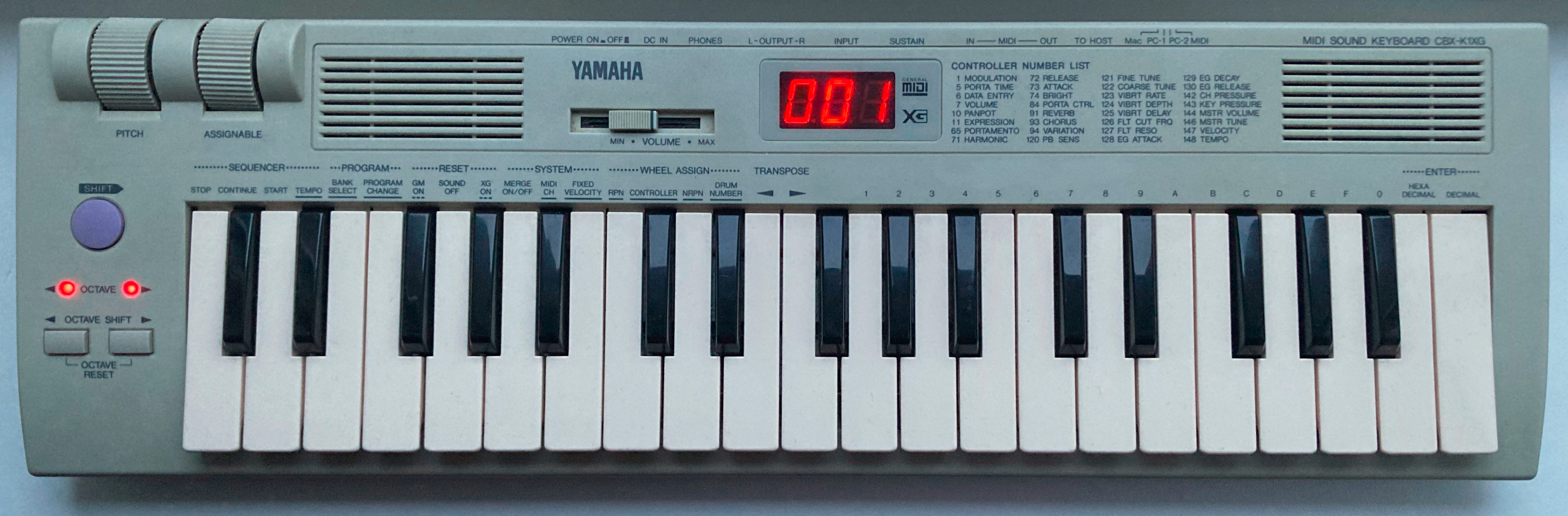 A photo of a “Yamaha MIDI Sound Keyboard CBX-K1XG”. This is a small, battery-powered musical keyboard with 37 mini-keys, and its enclosure is made from the same dull grey/beige plastic typical of 1990's computer equipment. Visible from this top-down perspective, it features a “Pitch” wheel, an “Assignable” wheel, a large purple “Shift” button, two red “Octave” LEDs, two “Octave Shift” buttons, a volume slider, a pair of speaker grilles, and a red-on-black three-digit seven-segment LED display showing the patch number “001”. It also features a lot of printed text on the enclosure referencing many different kinds of highly technical MIDI controller functionality, and labels for many kinds of connections that can be made at the rear of the unit.
