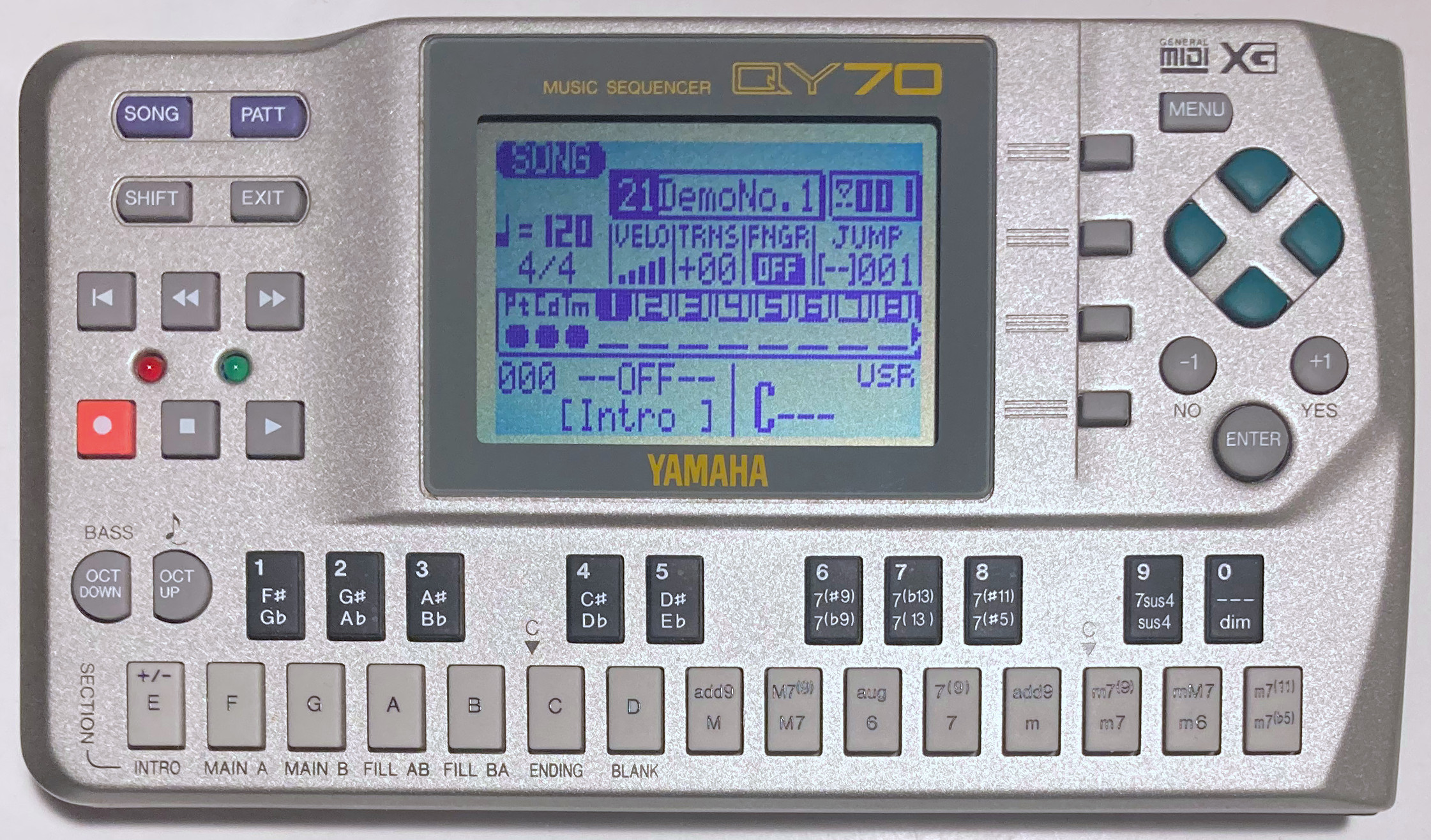 A photo of a “Yamaha QY70 music sequencer”. This is the smallest of the devices seen so far, smaller even than the SC-7, and perhaps similar in size to a VHS tape. It is a portable, battery-powered device that has a curved, angled, incredibly 1990's sparkling silver plastic design. It features a large, non-backlit dot-matrix LCD, on which we can see among other things that this is the “SONG” view, that song #21 “DemoNo.1” is selected, that the tempo is 120bpm, that the song is in 4/4 time, that the current pattern is #000 “Intro” and that the current chord is “C---”. There are also various settings for keyboard input such as velocity, and several sequencer tracks including special ones for Patterns, Chords and Tempo (abbreviated Pt, Cd and Tm). Outside the screen, there are 25 tiny rubber buttons arranged in a musical keyboard layout, which are labelled for entering numbers, selecting chord roots notes and chord types. There are also small rubber buttons in a D-pad shape, a menu button, play/stop/rewind/record controls, and “Song” and Pattern (abbreviated ”Patt”) buttons, among otherss.